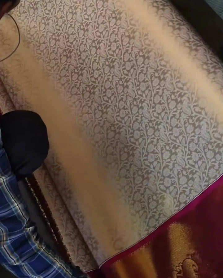 A close-up of a patterned fabric or saree with a beige and brown floral vine motif, a deep magenta border, and a gold zari ornamental section at the bottom.