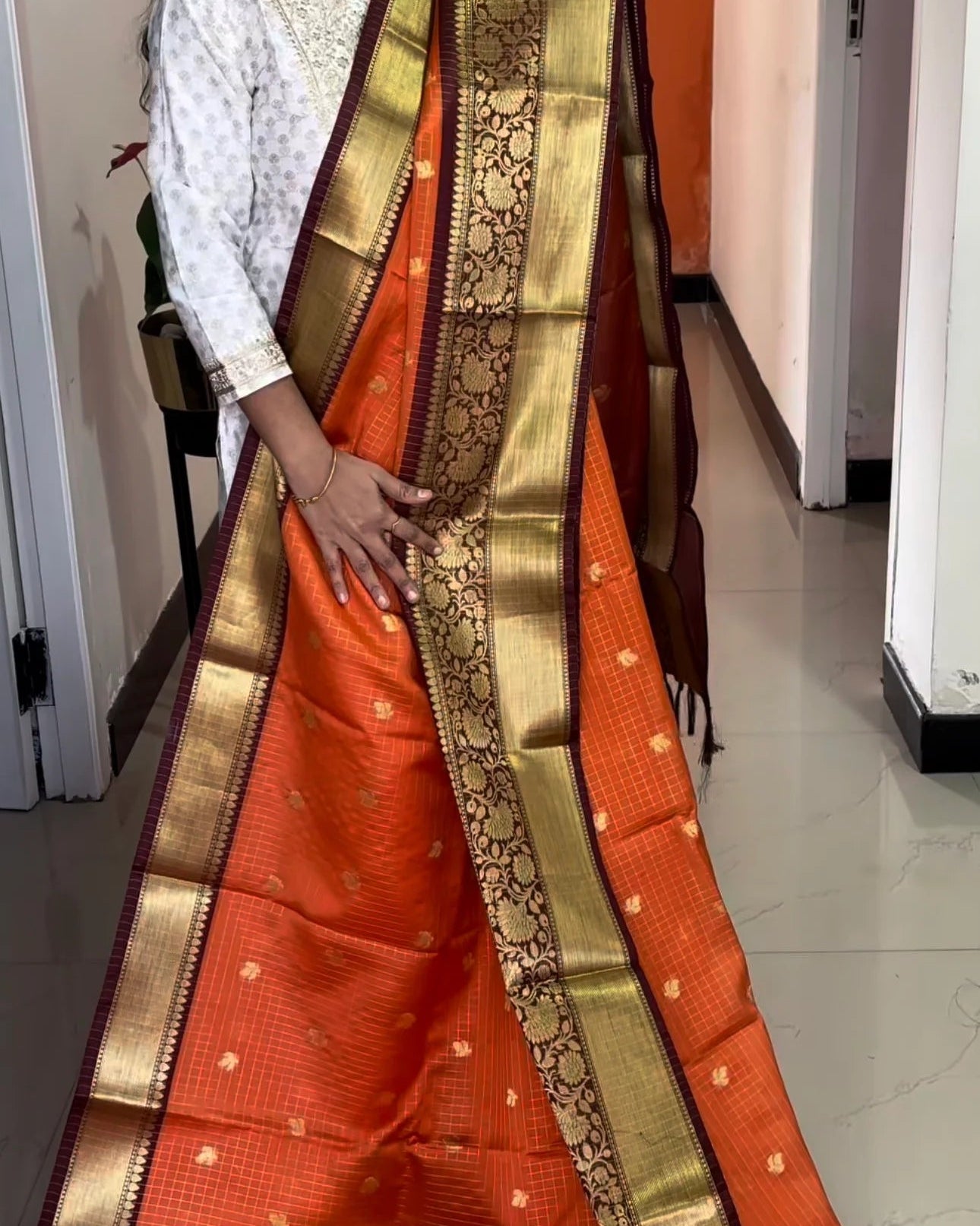 Person wearing an orange saree with a gold border indoors.
