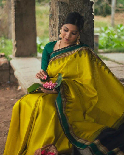Woman in a yellow saree with green border sitting outdoors.