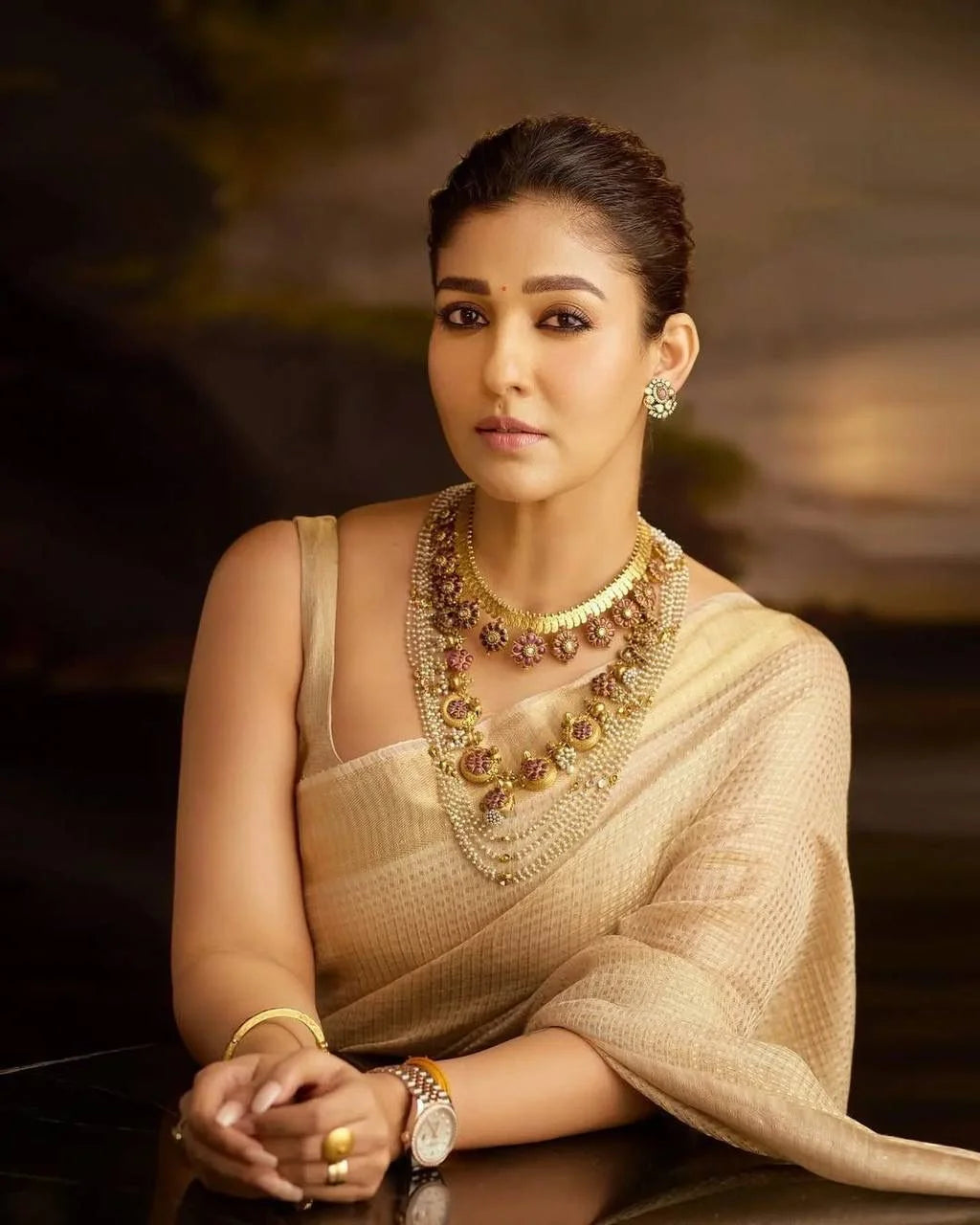 Woman wearing a beige saree with gold jewelry against a dark background